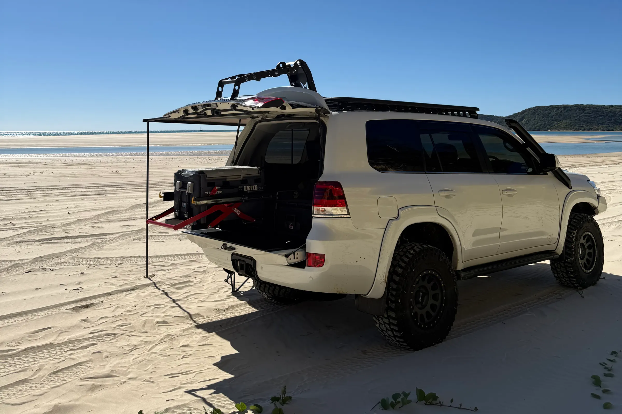 A white 200 Series on the beach, rear door up and a fridge on a drop slide, awning pole in the sand.