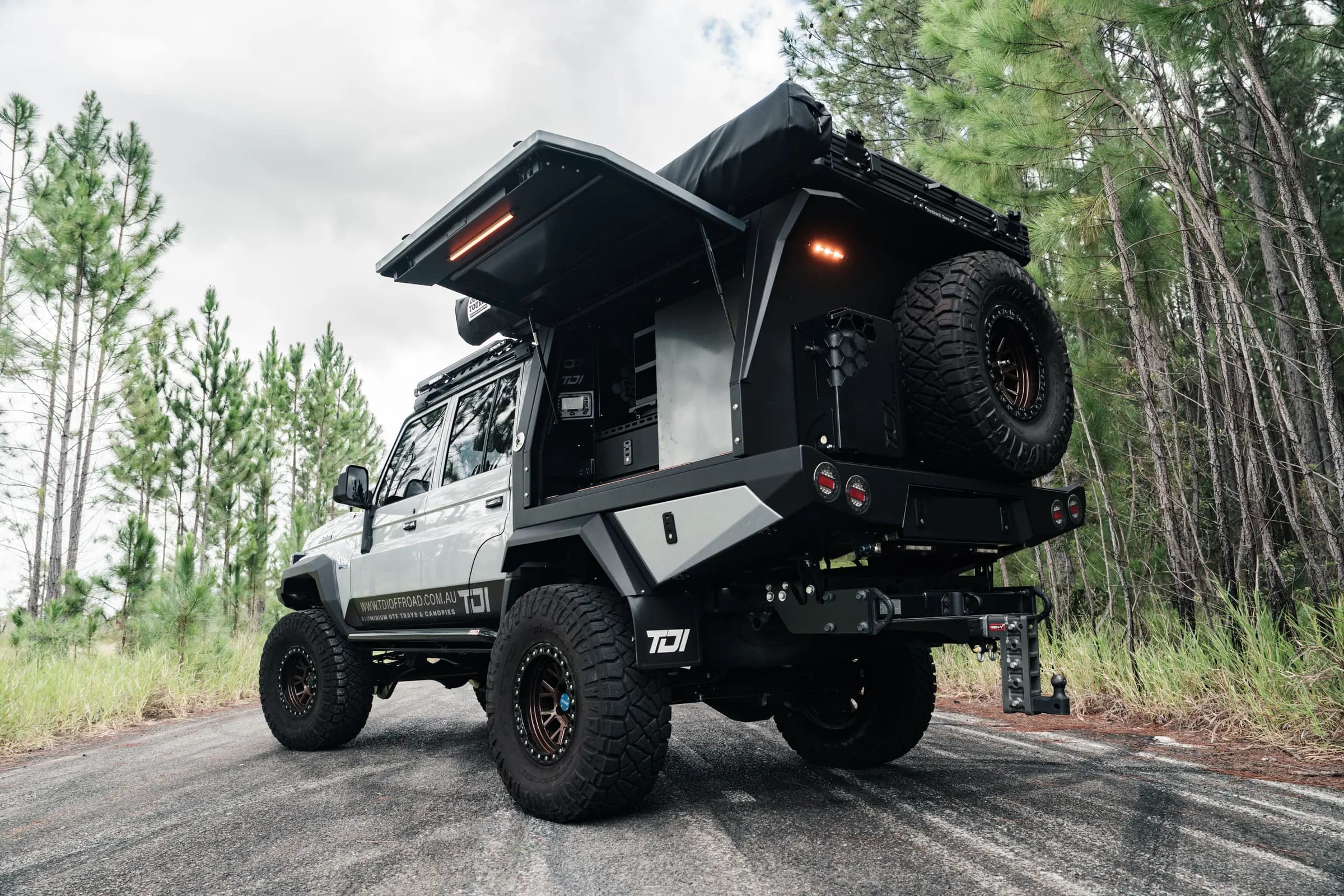 A lifted 79 Series dual cab with a TDI tray and canopy, gull-wing door up, on a road through a pine forest.