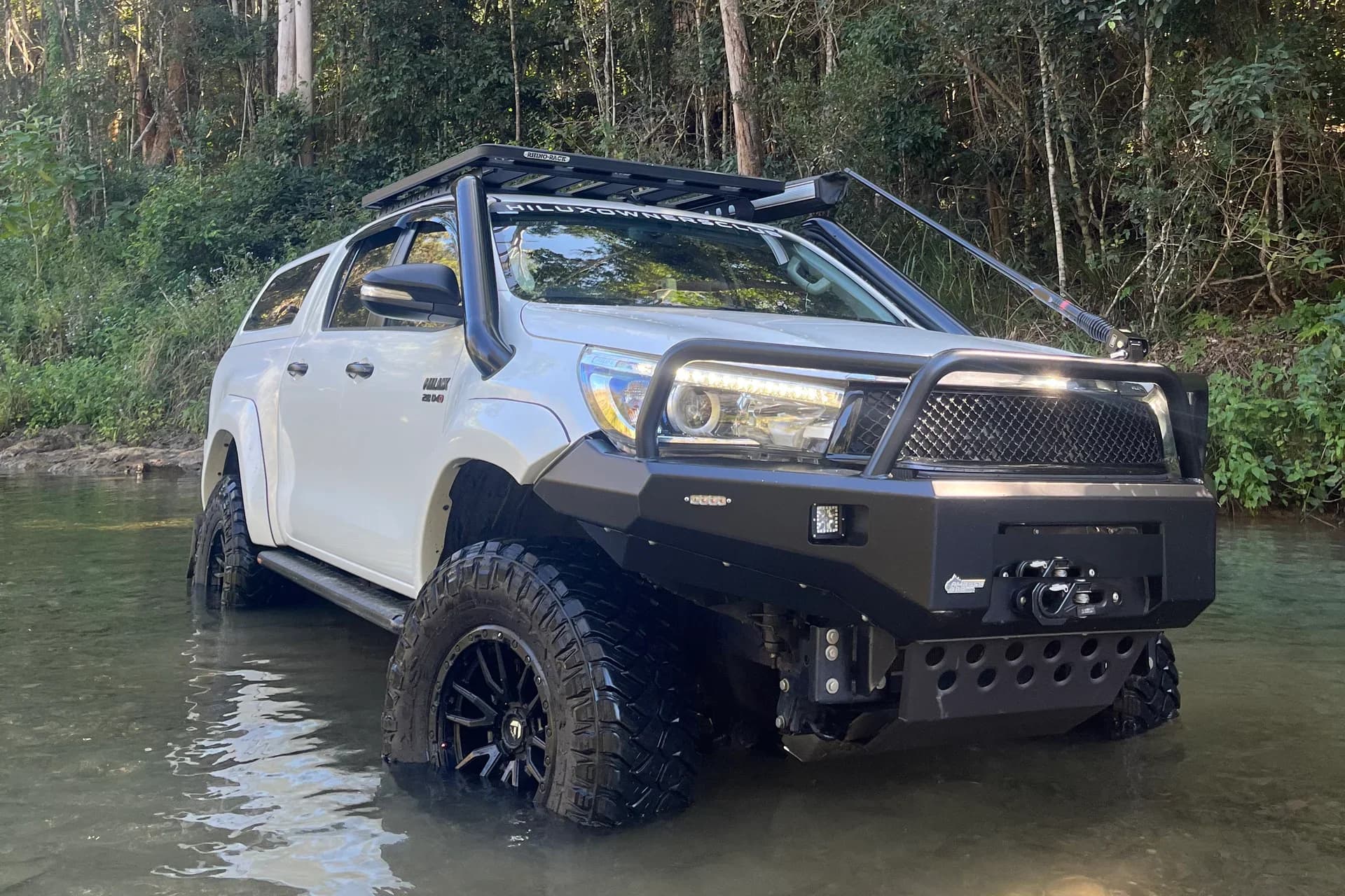 A white Hilux with a bull bar and roof rack parked in a shallow creek, rainforest behind.