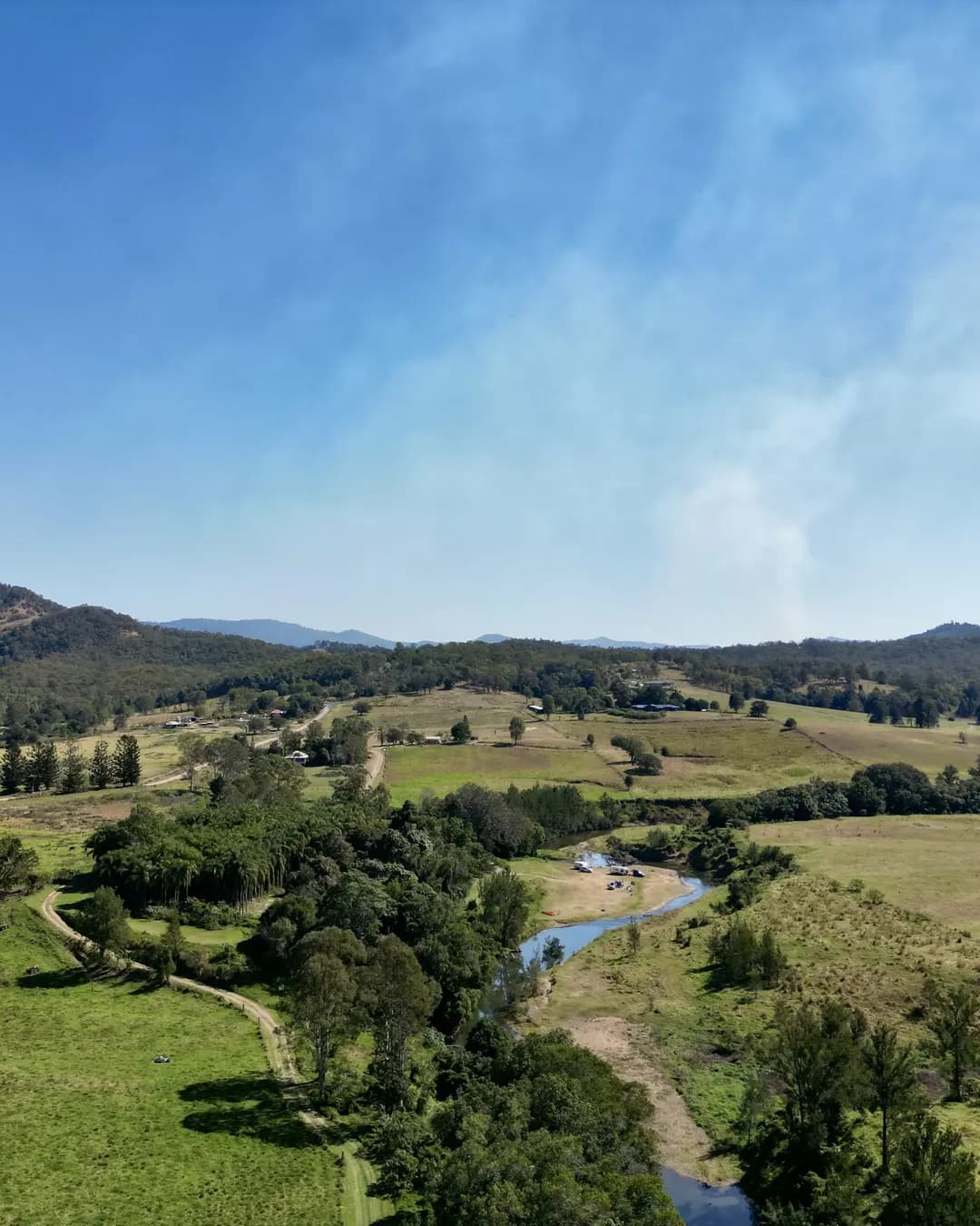 Drone view over green river flats, a creek winding through the trees and ranges behind.
