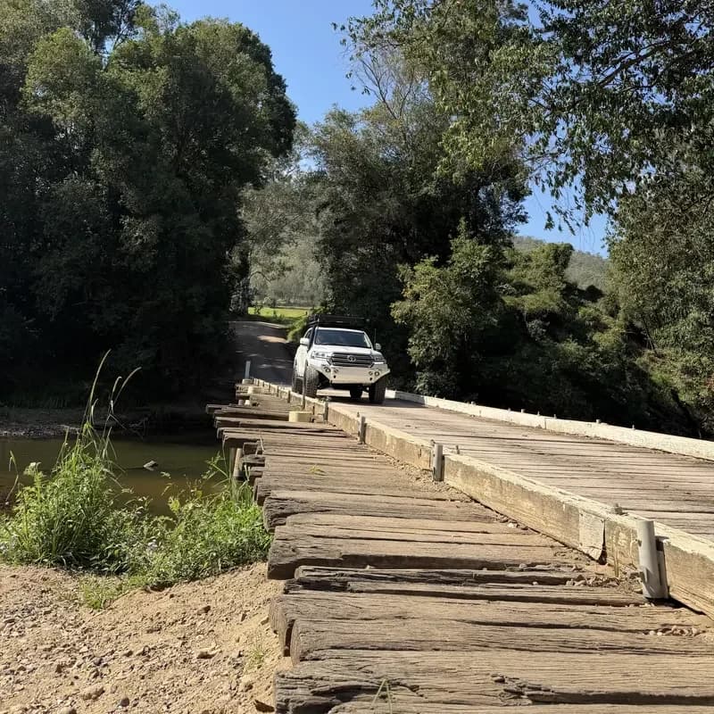 A 200 Series rolling onto an old timber bridge between the trees.