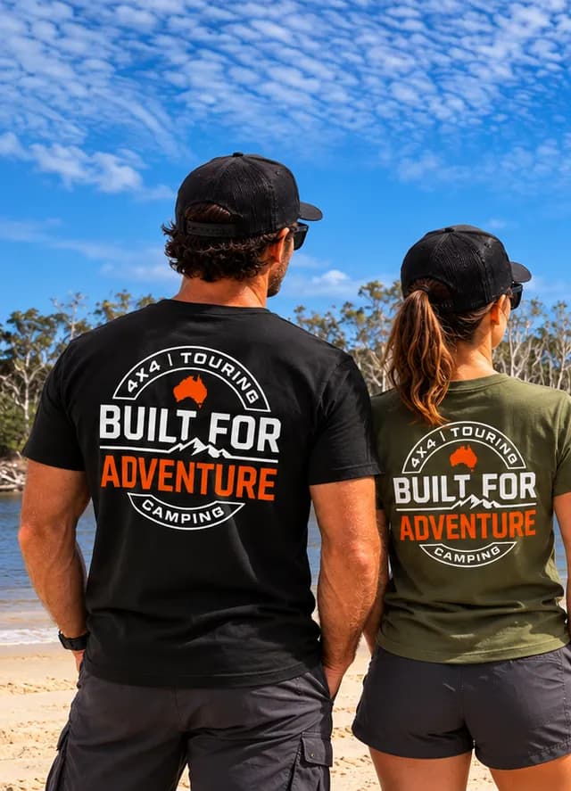 Two people on a beach in Built for Adventure tees, seen from behind